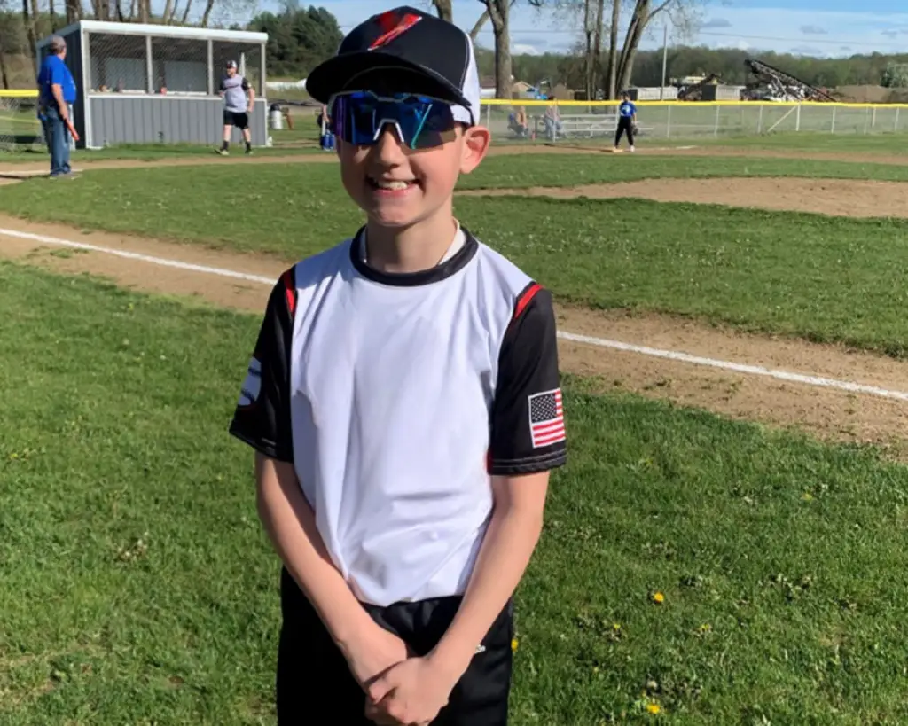Maxton, a patient with a congenital heart defect called tricuspid atresia standing in his baseball uniform in front of a baseball field