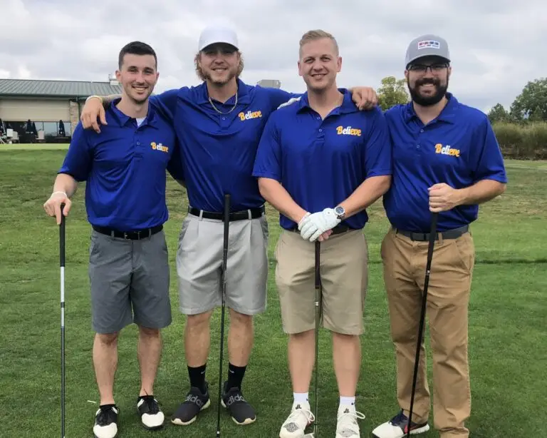 A group of four men standing on a golf course, holding golf clubs, and wearing matching blue polo shirts