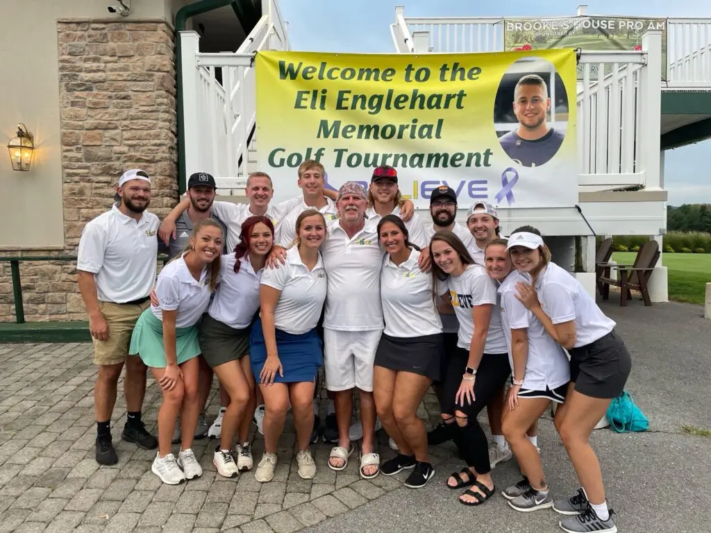 A group of people standing in front of a banner that reads "welcome to the Eli Englehart Memorial Golf Tournament." They are wearing matching white polo shirts.