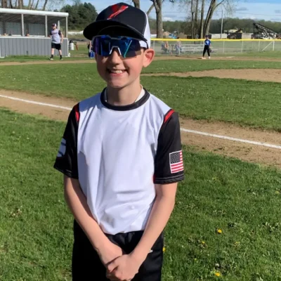 Maxton, a patient with a congenital heart defect called tricuspid atresia standing in his baseball uniform in front of a baseball field
