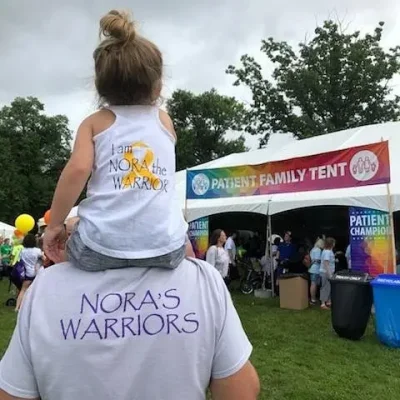 A girl on her dad's shoulders facing away from the camera. Their shirts say "Nora's Warriors"